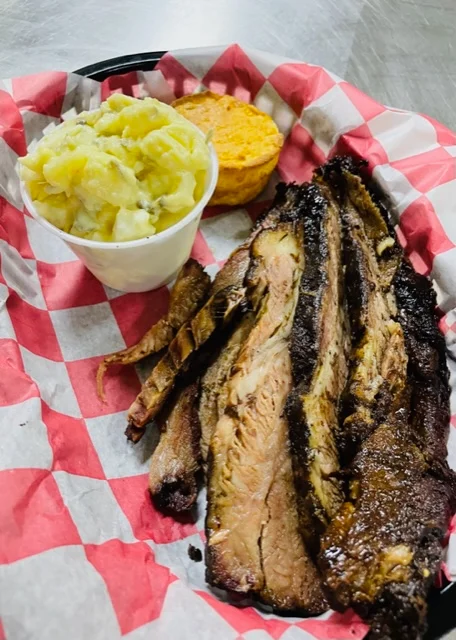 A serving of smoked brisket with a side of mashed potatoes and a biscuit, presented in a red checkered paper basket.