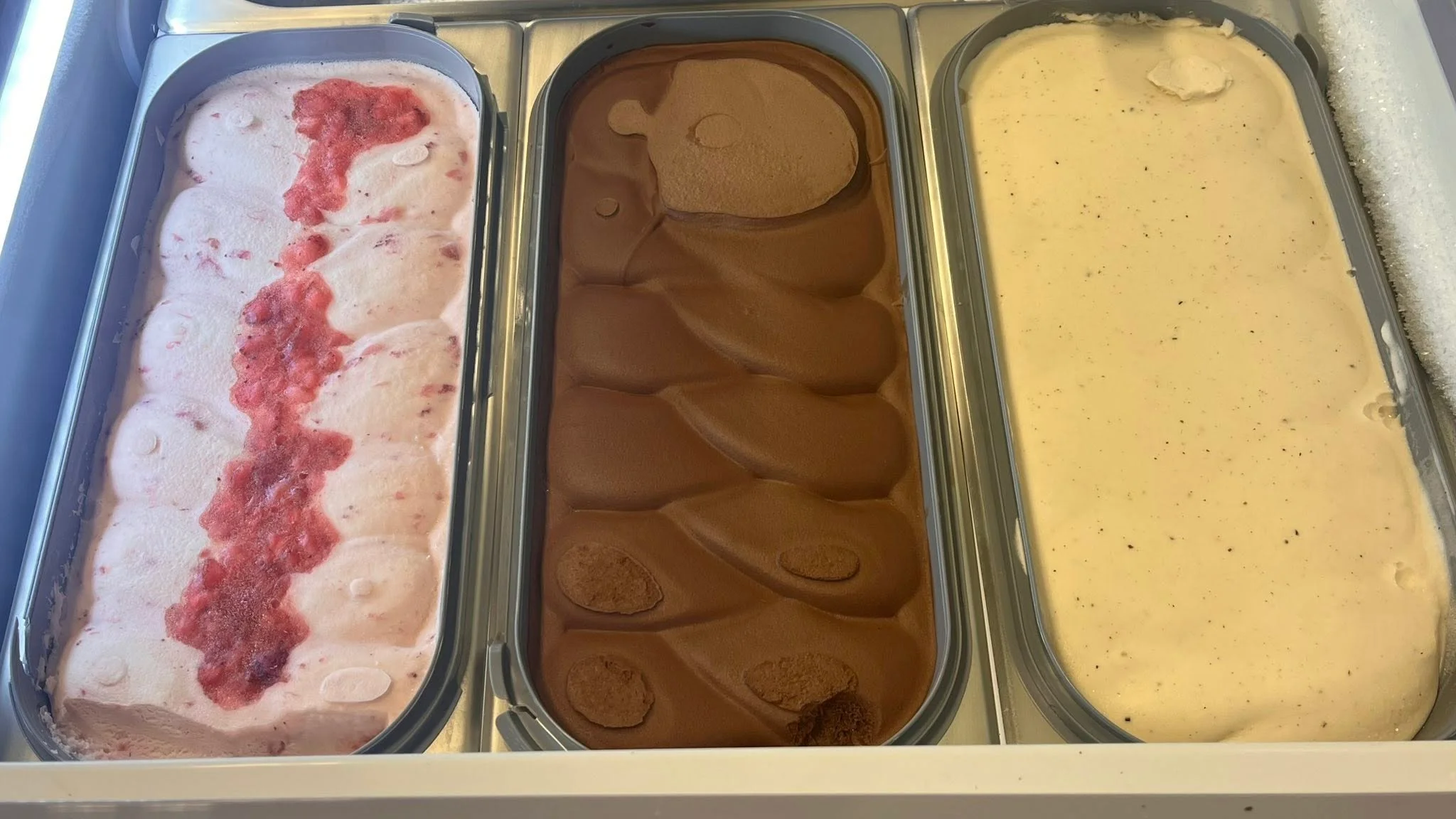 Three containers of gelato featuring different flavors: a strawberry swirl, a rich chocolate, and a vanilla-like flavor. Each container showcases a smooth texture.