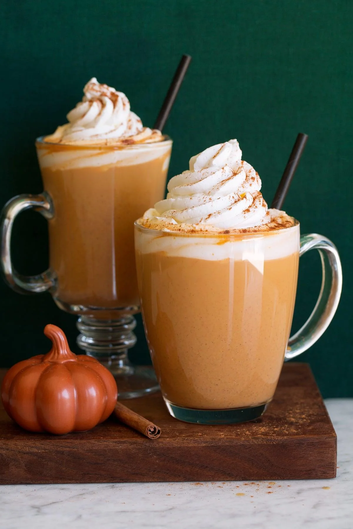 Two cups of creamy pumpkin spice drink topped with whipped cream and sprinkled with cinnamon. A small pumpkin and a cinnamon stick are nearby.