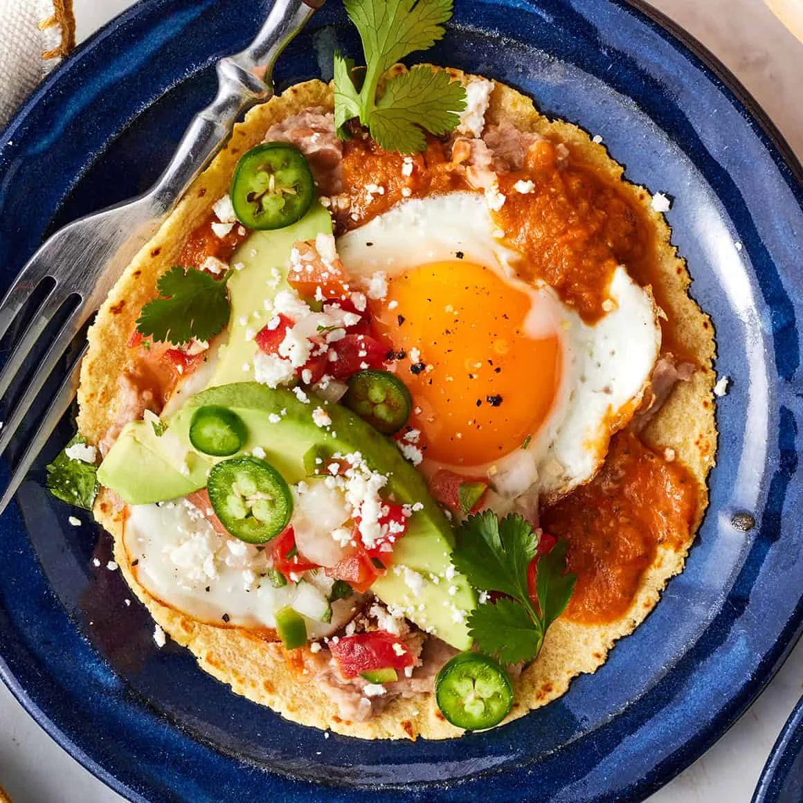 A colorful breakfast plate featuring a taco topped with a fried egg, avocado slices, and fresh vegetables, garnished with cilantro.