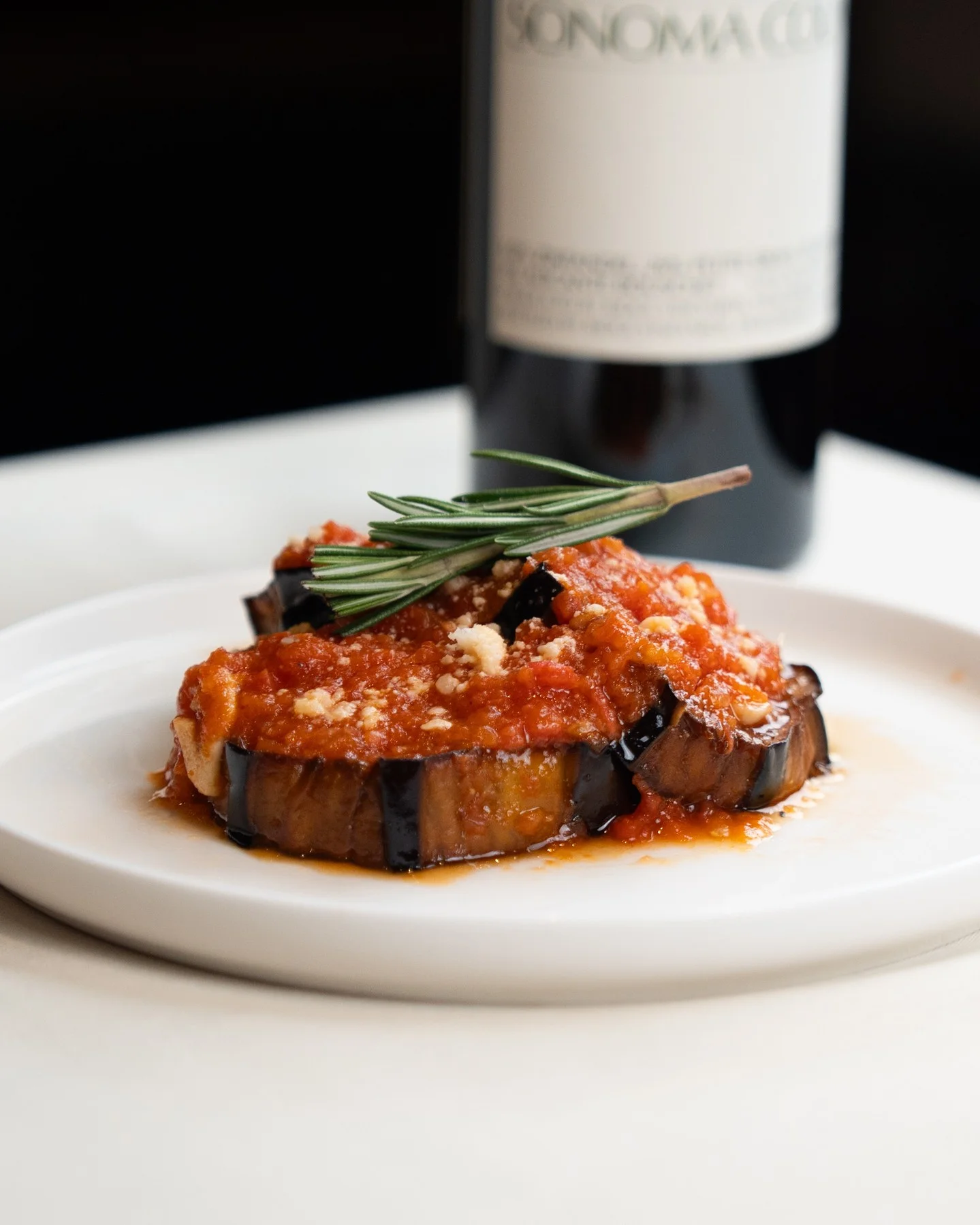 A plate of roasted eggplant topped with tomato sauce and a sprinkle of cheese, garnished with a sprig of rosemary.