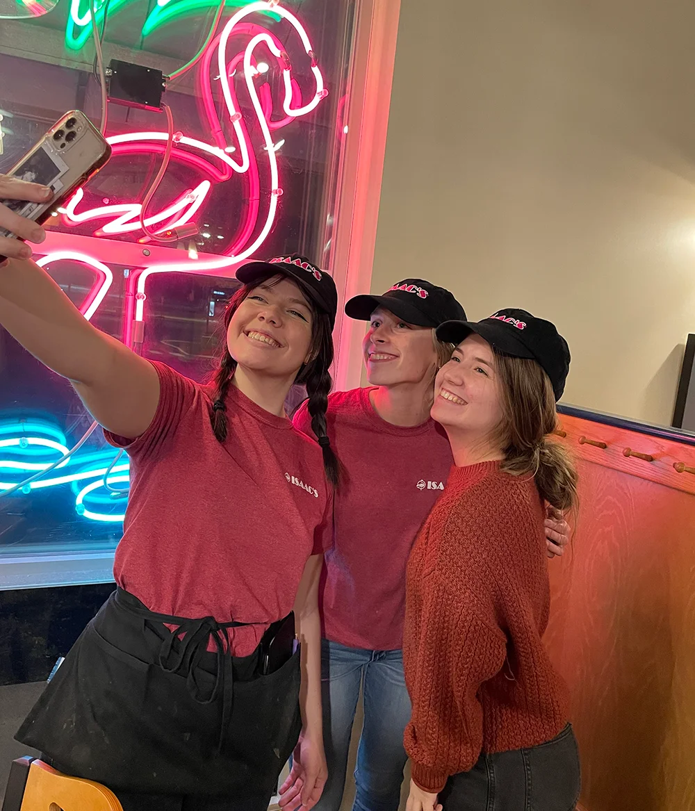 Three friends in red Isaac's shirts capturing a fun selfie against a bright neon flamingo sign backdrop.