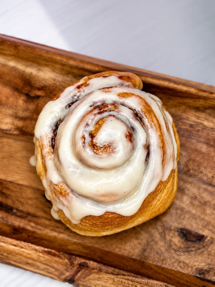 A freshly baked cinnamon roll topped with cream icing, displayed on a wooden plate.