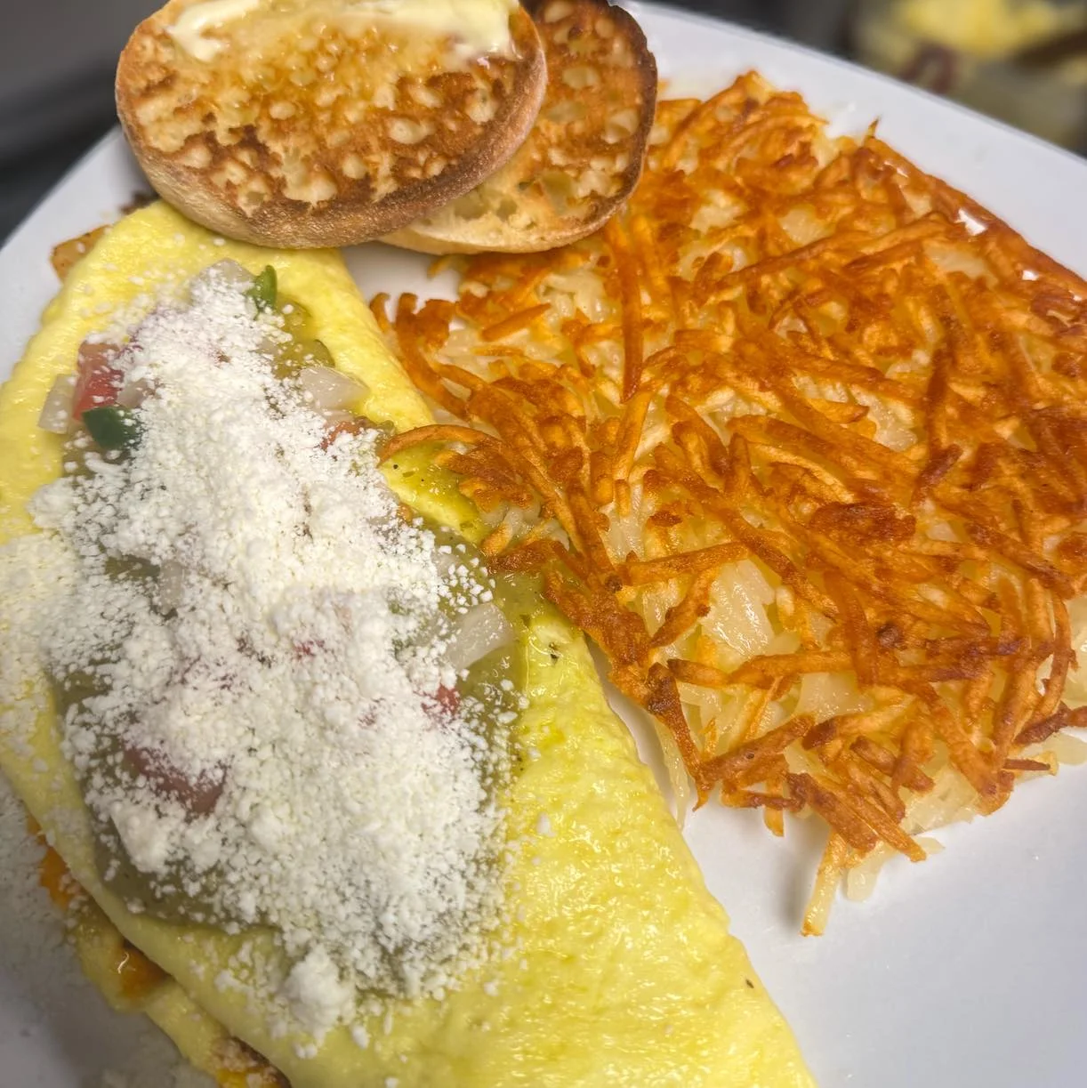 A delicious breakfast plate featuring an omelet filled with vegetables, topped with cheese, accompanied by crispy hash browns and toasted English muffin halves.