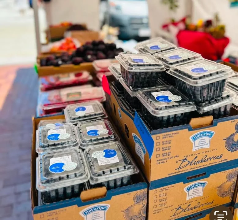 A variety of fresh blueberries packaged in plastic containers are displayed at a market stand, with other fruits in the background.