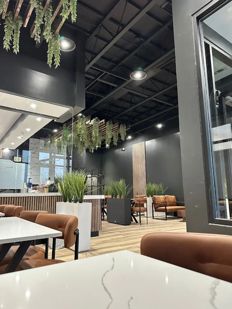 An interior view of a modern dining space featuring stylish seating, greenery, and contemporary decor. The space includes wooden flooring and a combination of light and dark color palettes.