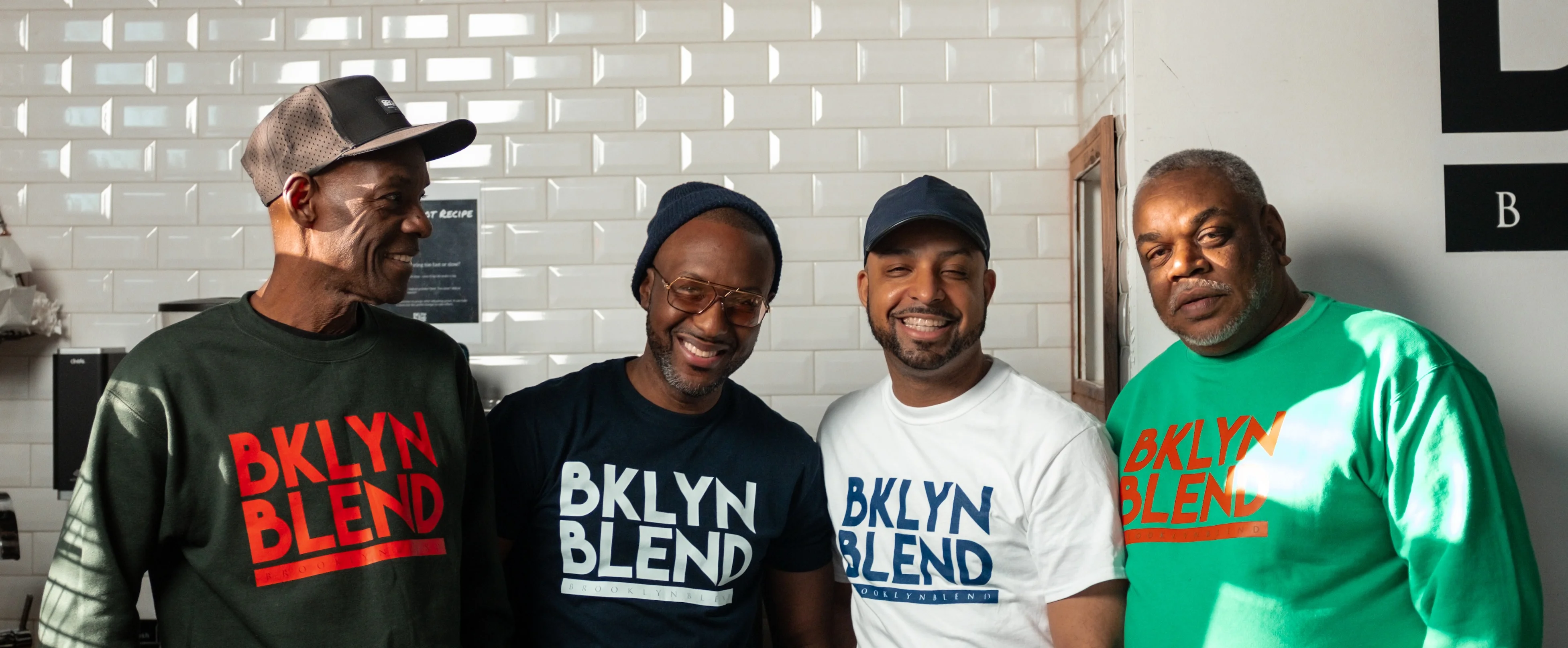 Four smiling men stand side by side wearing "BKLYN BLEND" shirts. The setting has white tiled walls, creating a warm and friendly atmosphere.