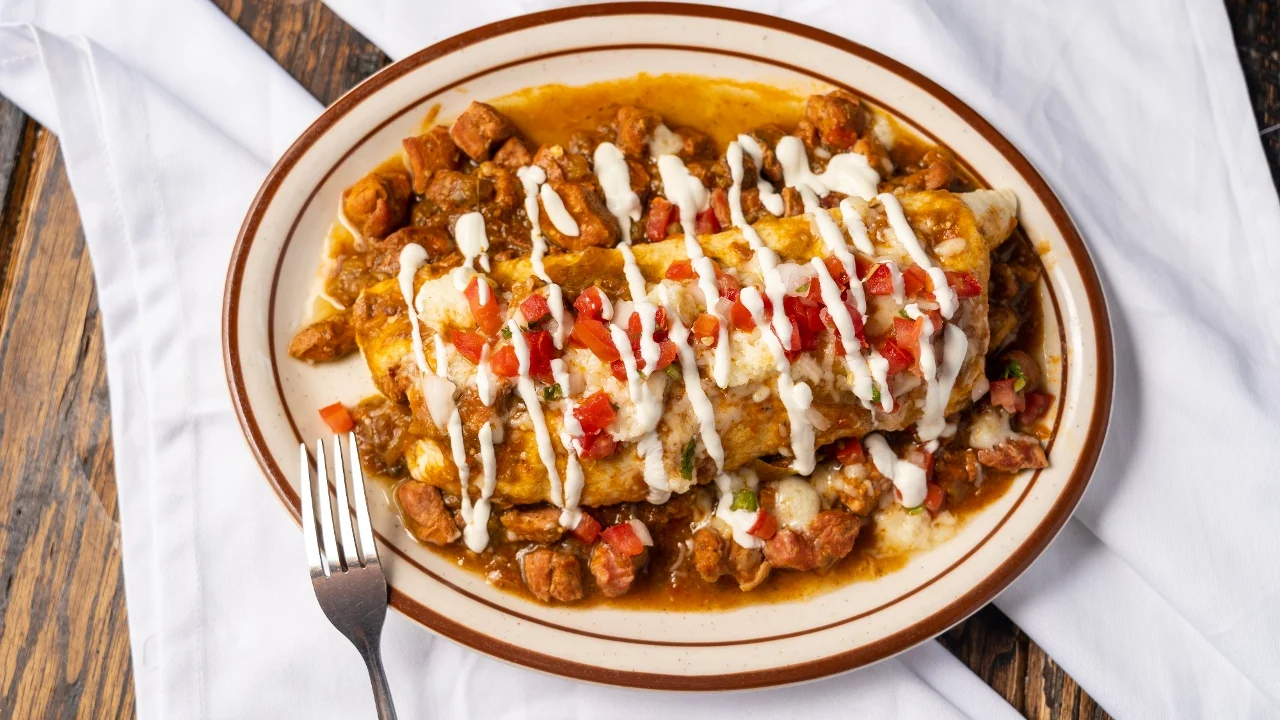 A plate with a large burrito covered in sauce, garnished with sour cream and diced tomatoes, alongside smaller pieces of meat.