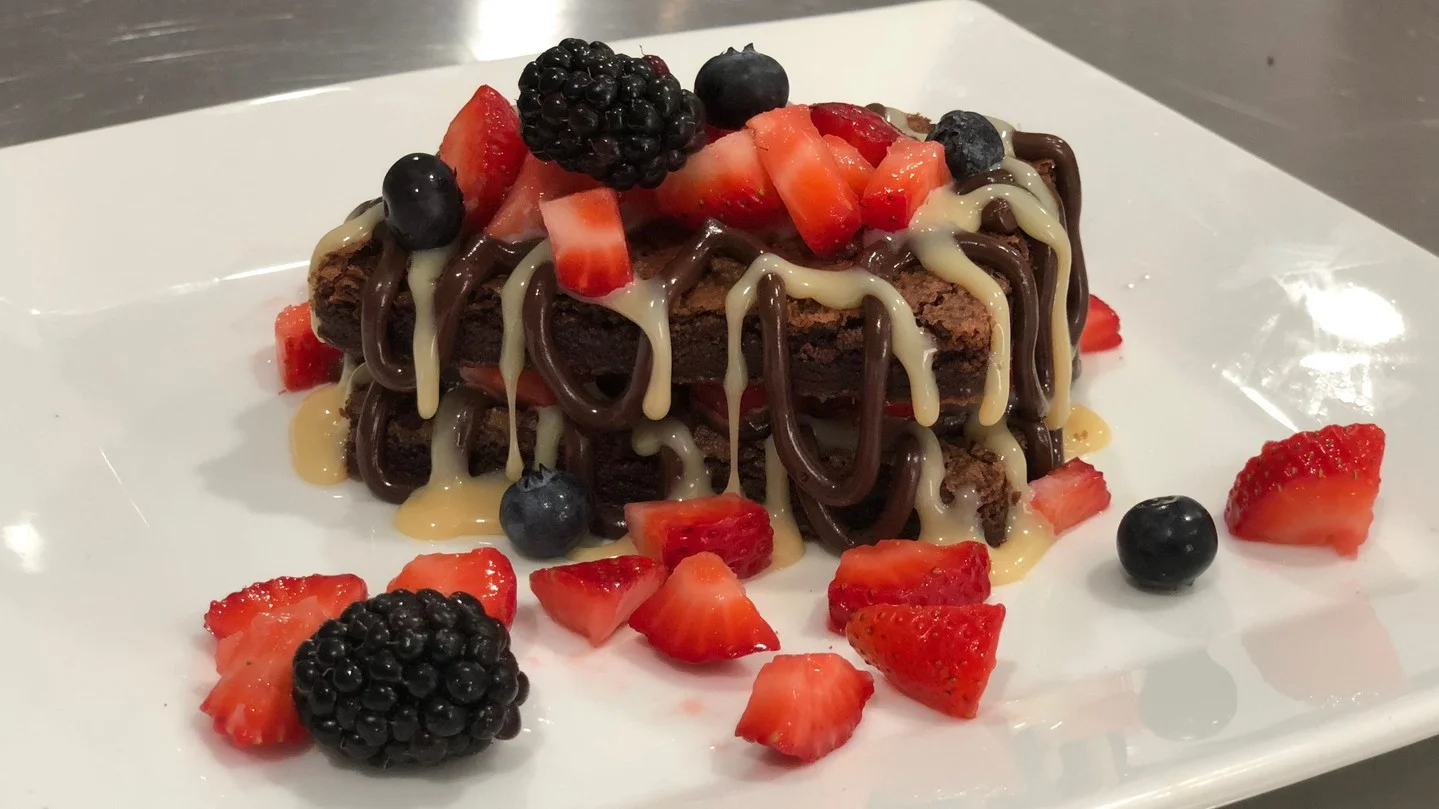 A dessert featuring a brownie slice drizzled with chocolate and cream, topped with fresh berries.