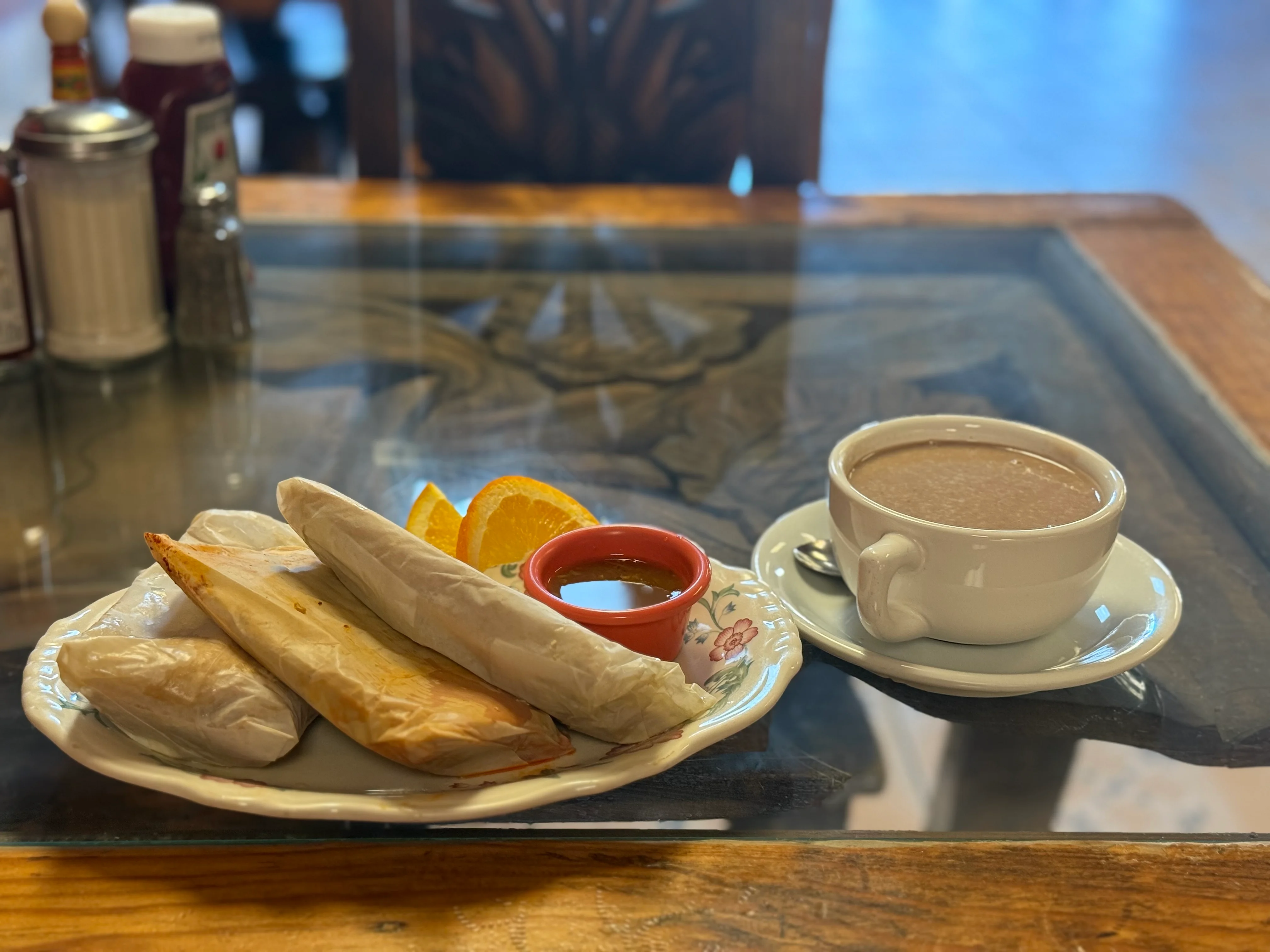 A plate with three wrapped items, orange slices, and a bowl of salsa, alongside a cup of hot chocolate on a wooden table.