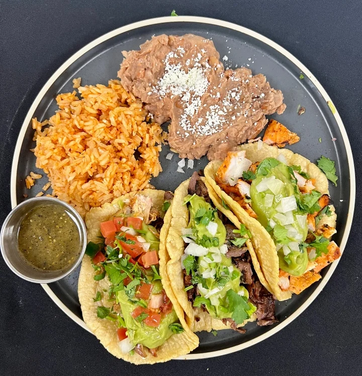 Three enchiladas garnished with cheese, tomatoes, and sour cream, served with a side of guacamole on a black plate.