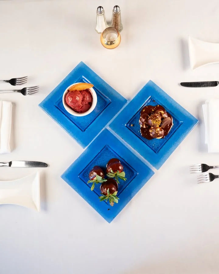 A trio of desserts served on blue plates, including a bowl of sorbet, a chocolate-covered dessert, and a plate with chocolate-covered treats, all elegantly arranged on a white tablecloth.