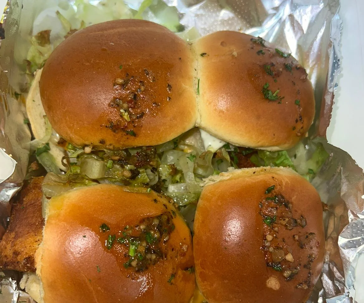 A serving of slider-style buns filled with flavorful ingredients, topped with a sprinkle of herbs and served in a foil-lined container.