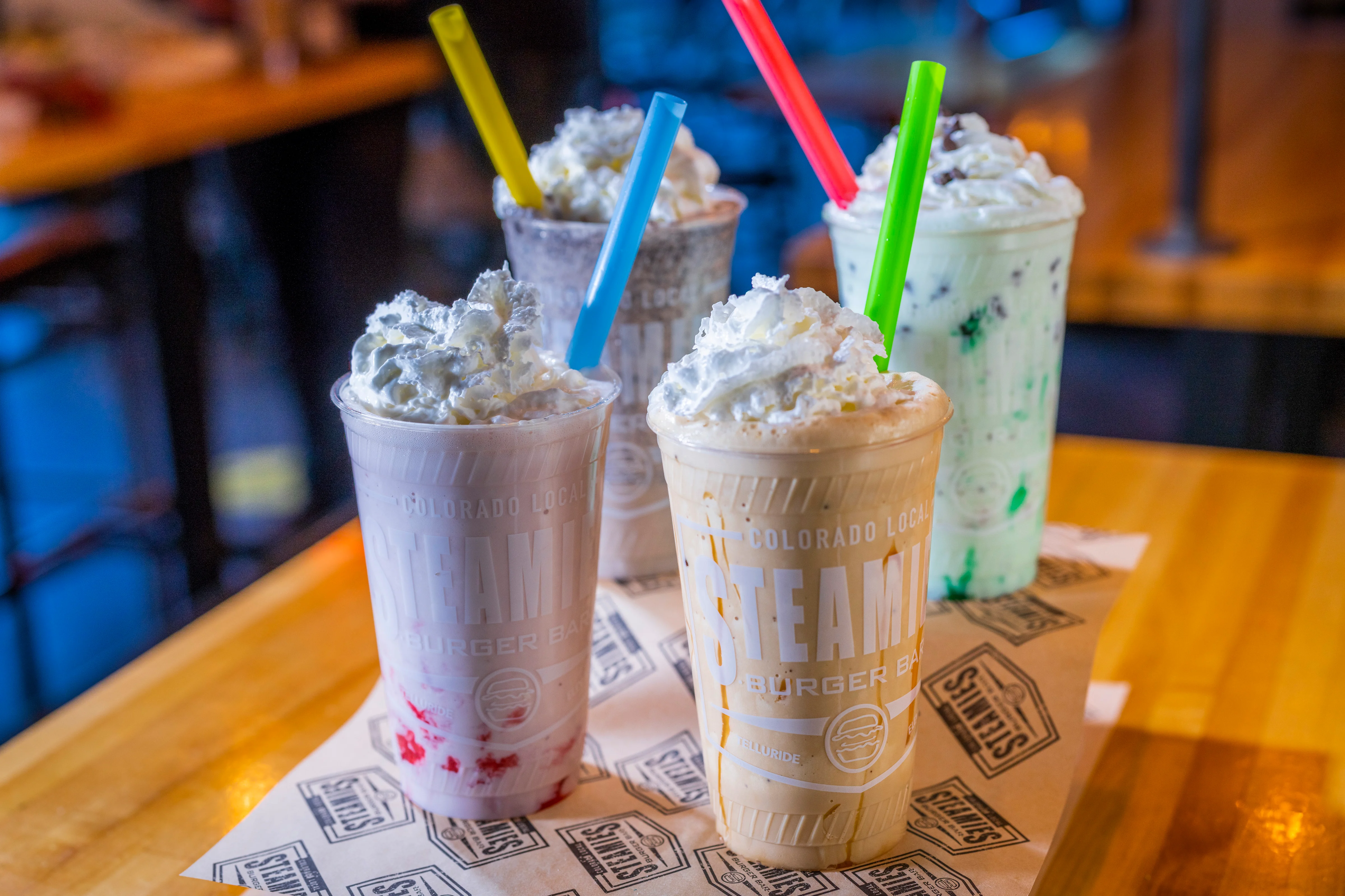 Four colorful milkshakes topped with whipped cream and served in clear cups with straws. Each shake features a different flavor, with vibrant colors and a creamy texture.