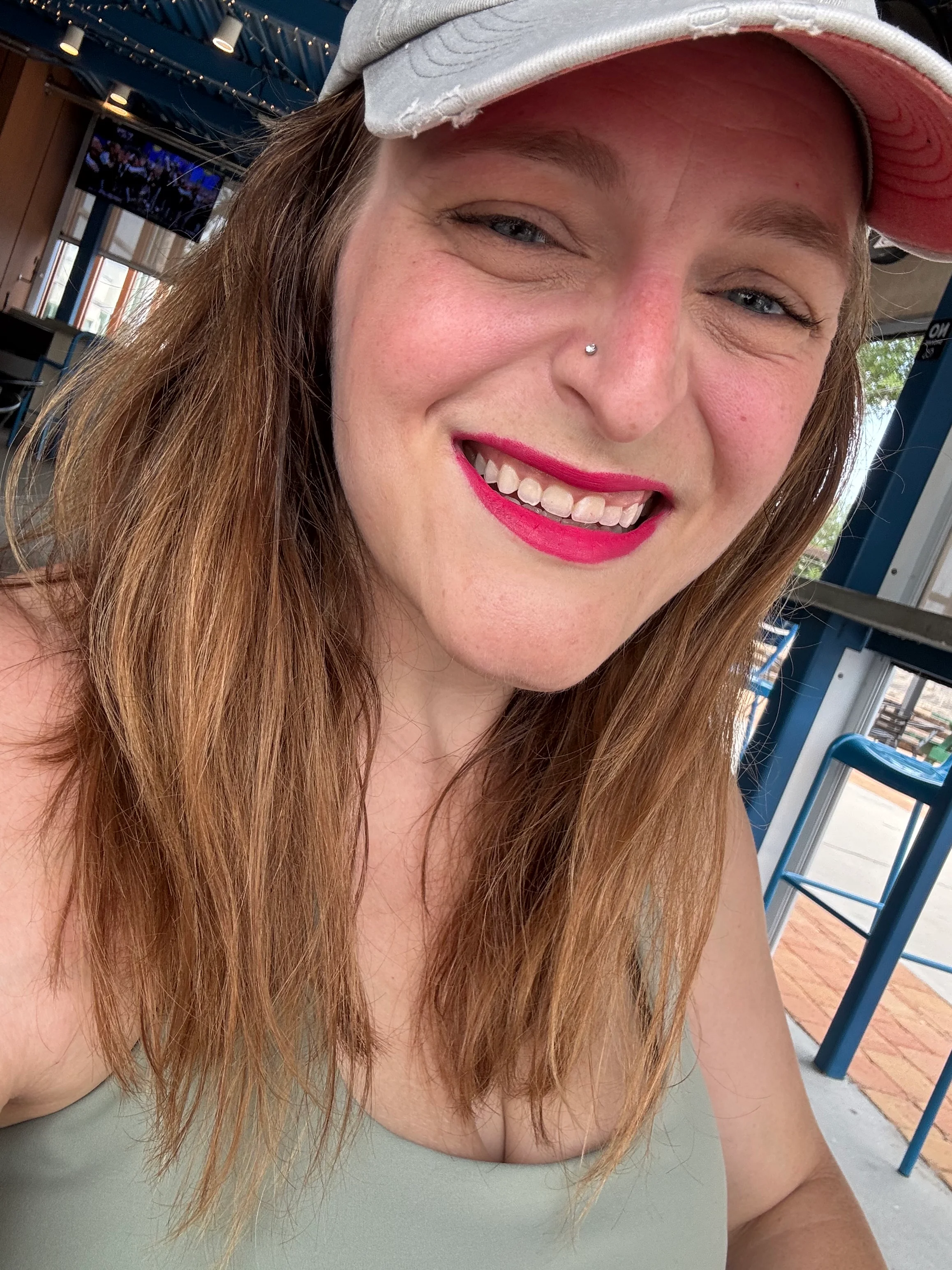 Smiling woman with long hair wearing a cap and bright lipstick in a casual setting.