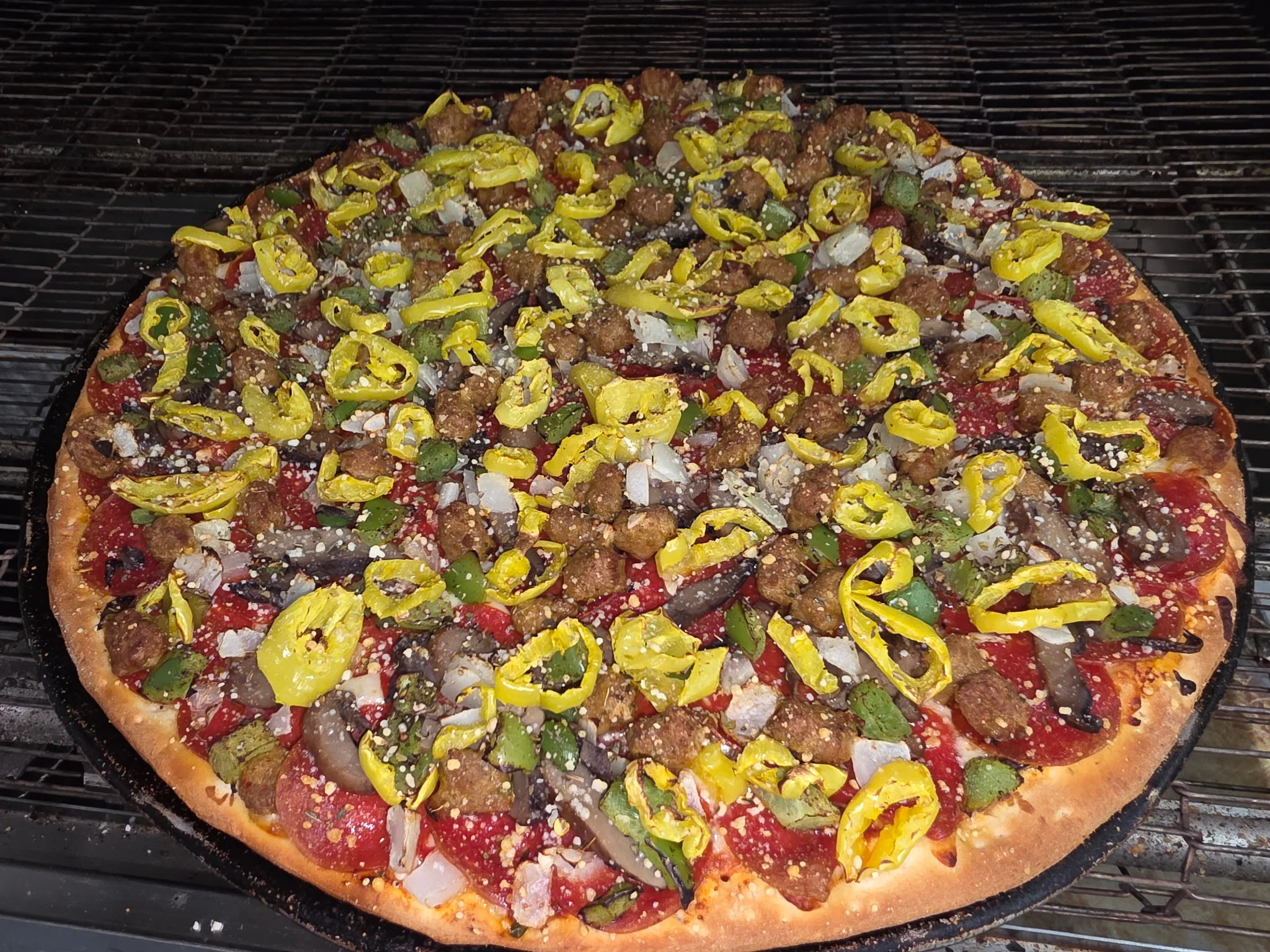 A freshly baked pizza topped with an array of meats, vegetables, and yellow peppers, sitting on a grill.