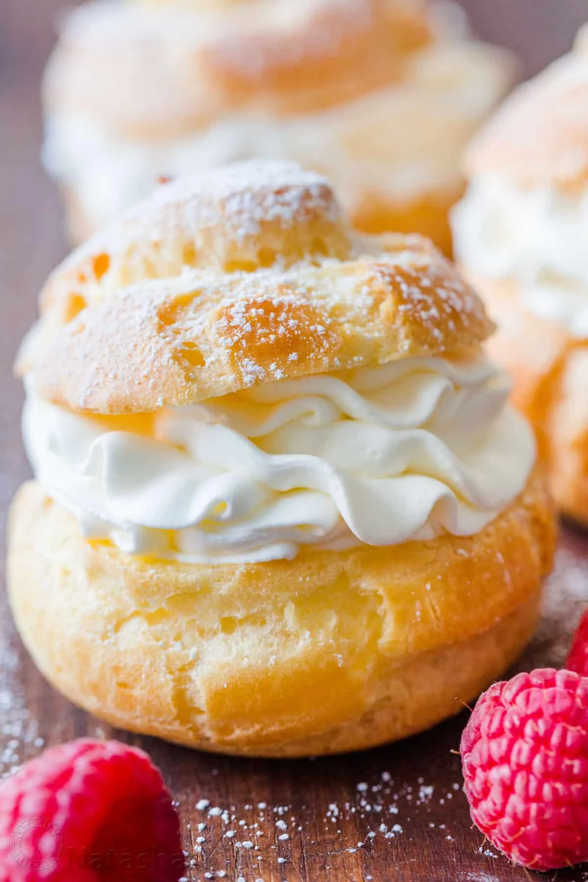 A close-up of a cream-filled pastry topped with powdered sugar, served alongside fresh raspberries on a wooden surface.