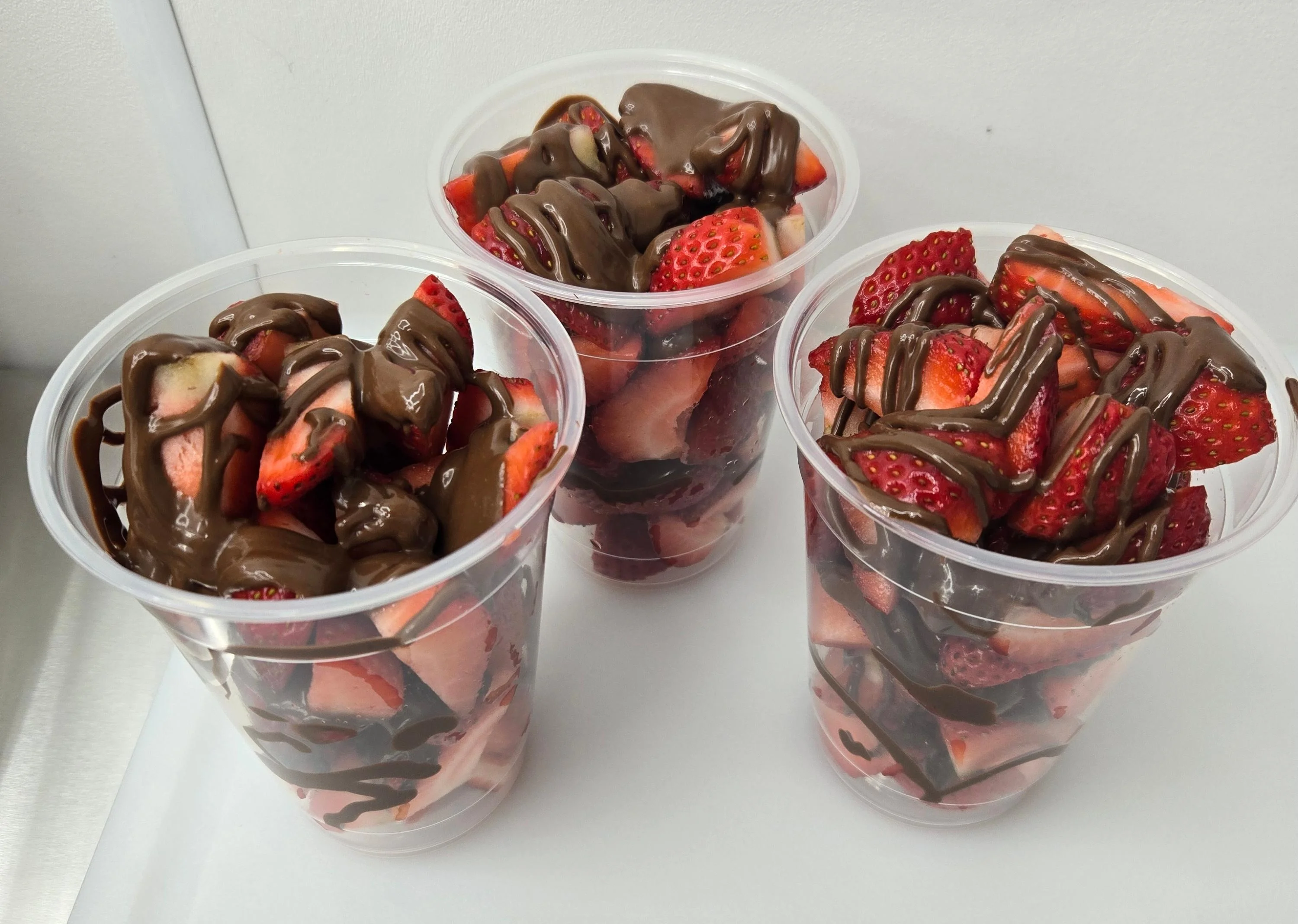 Three clear plastic cups filled with chopped strawberries drizzled in chocolate, showcasing a delicious dessert option.