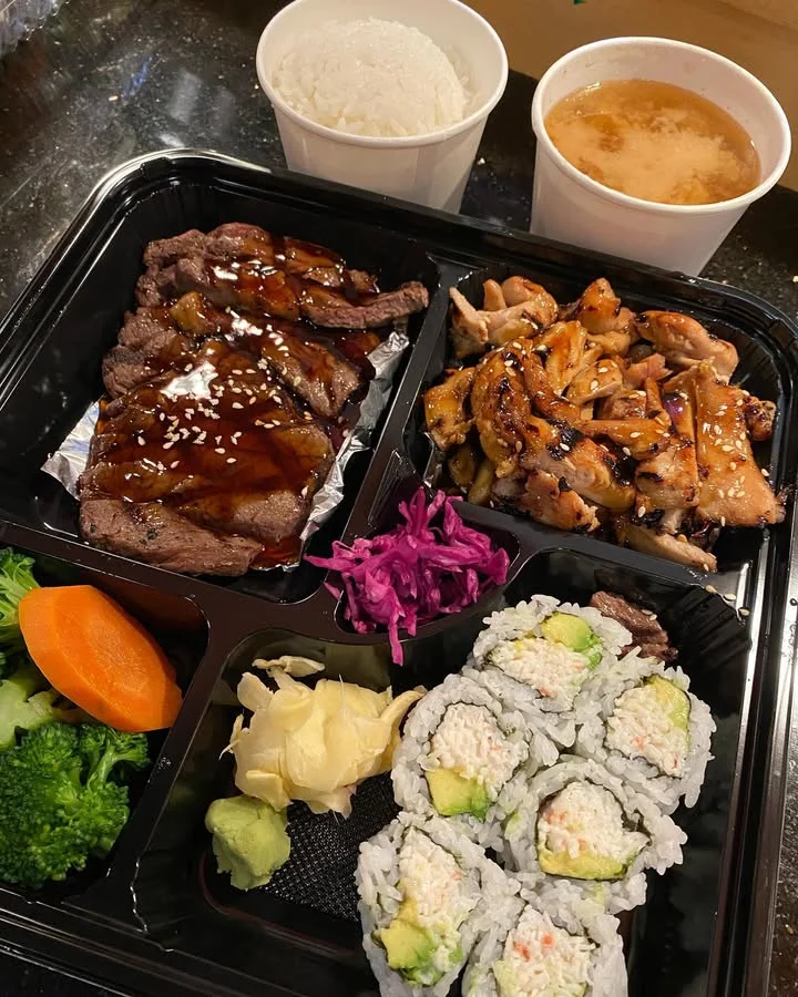 A food container with a variety of dishes including grilled beef, chicken, sushi rolls, mixed vegetables, and rice, accompanied by miso soup. The colorful presentation adds a vibrant touch.