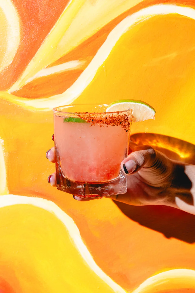 Hand holding a Mexican cocktail with lime and chili rim in front of a bright orange mural at Friducha in Brooklyn, NY.