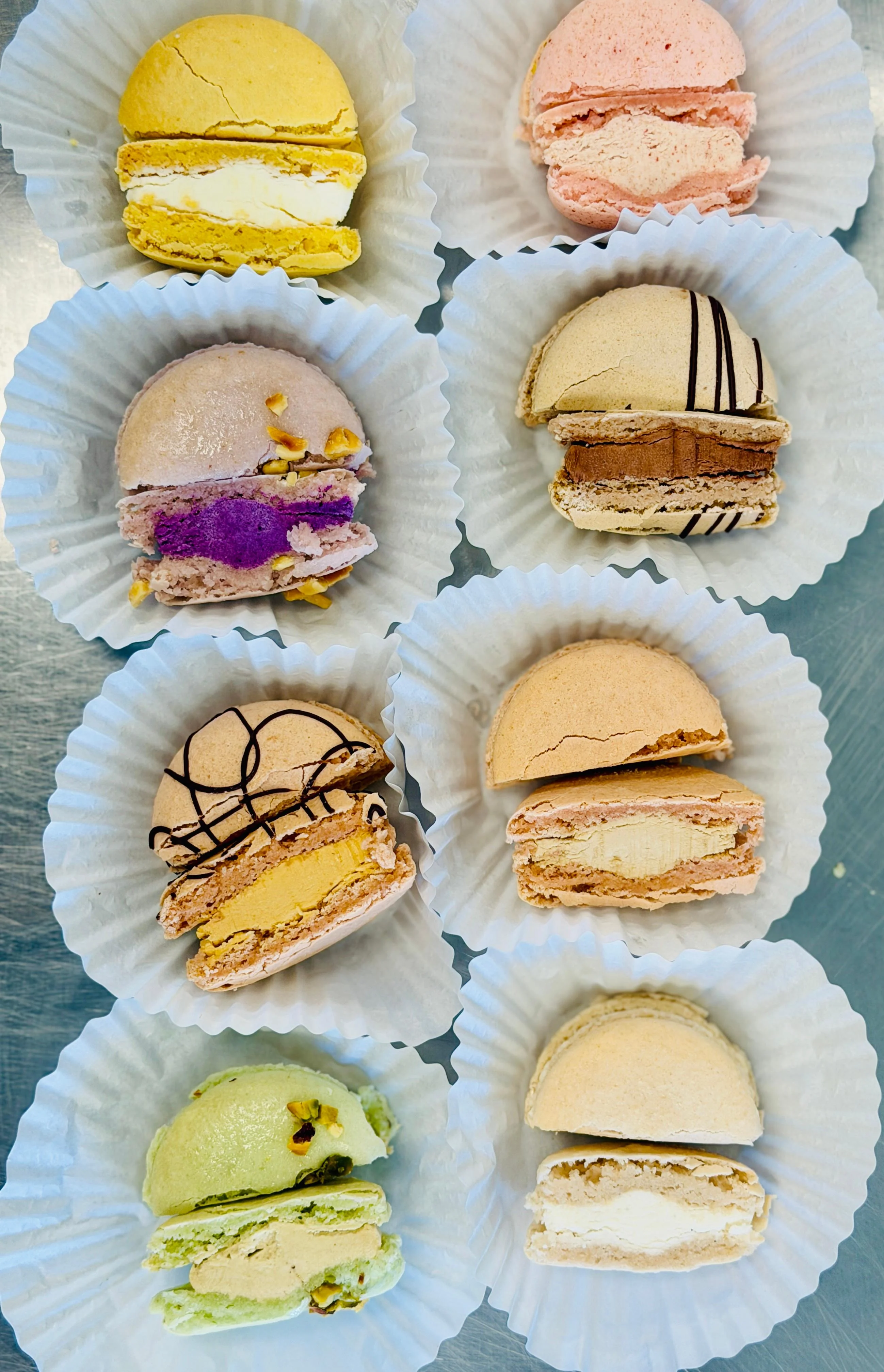An array of colorful macarons arranged neatly in white liners. The flavors include various fruits and chocolate, showcasing different colors and patterns.