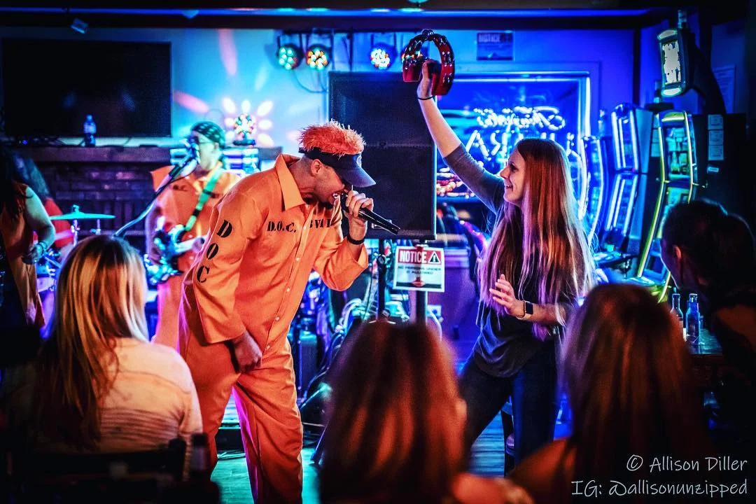 A lively performance with a musician in an orange outfit singing while a woman dances beside him, captured in a vibrant atmosphere filled with neon lights.