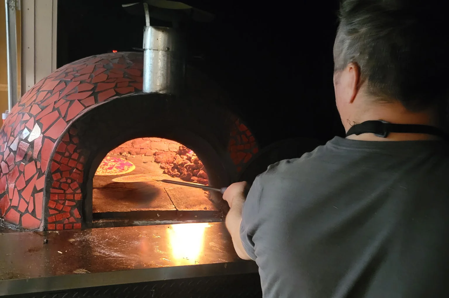 A person is pulling a freshly baked pizza from a wood-fired oven on a roasting cart. The oven is decorated with colorful tiles.