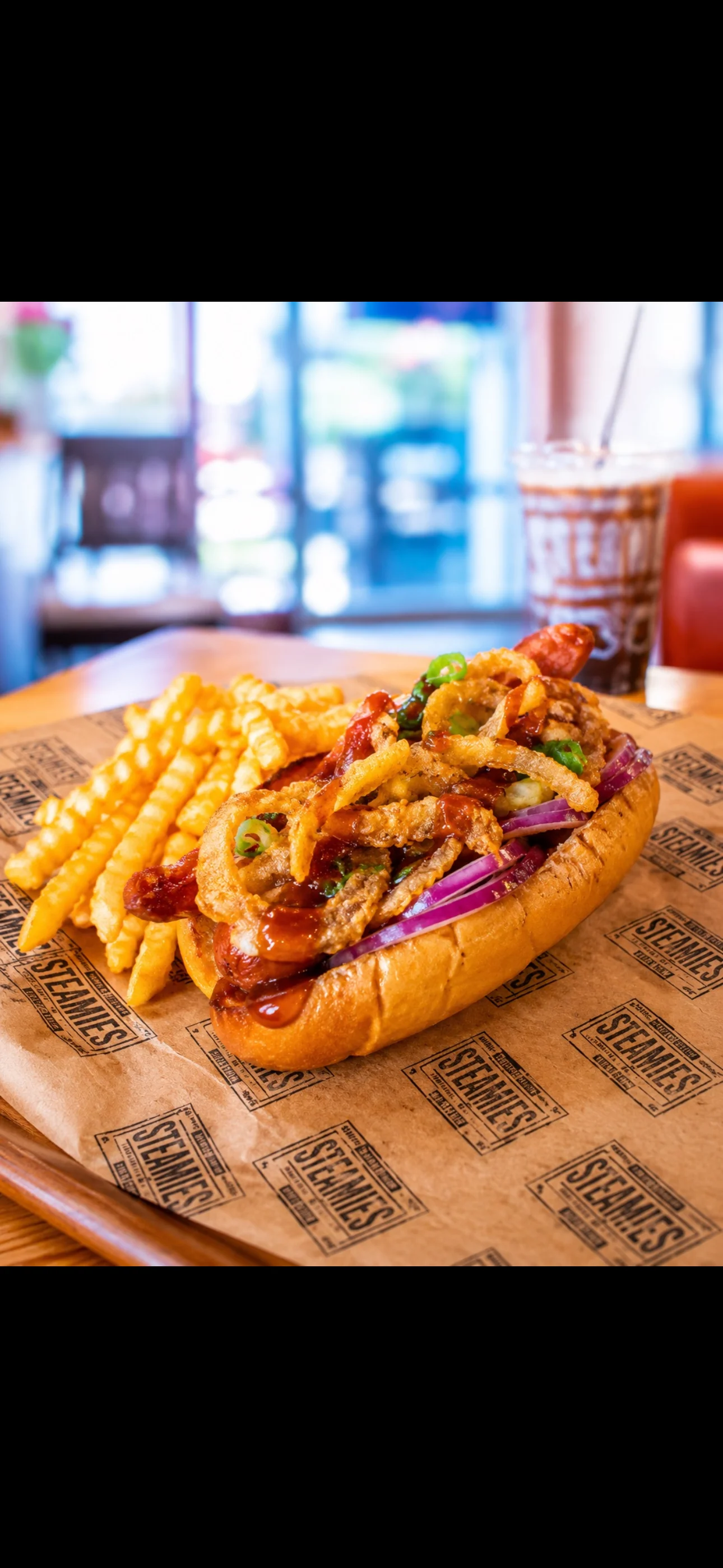 A delicious hot dog topped with sliced tomatoes, green onions, and a drizzle of sauce, served in a paper-lined basket.