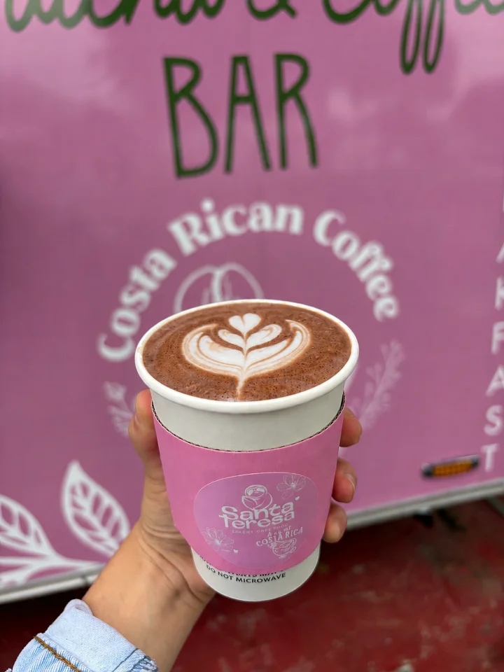 A cup of chocolate latte with a leaf design in foam, held in a hand. The cup has a pink sleeve and is in front of a pink sign.