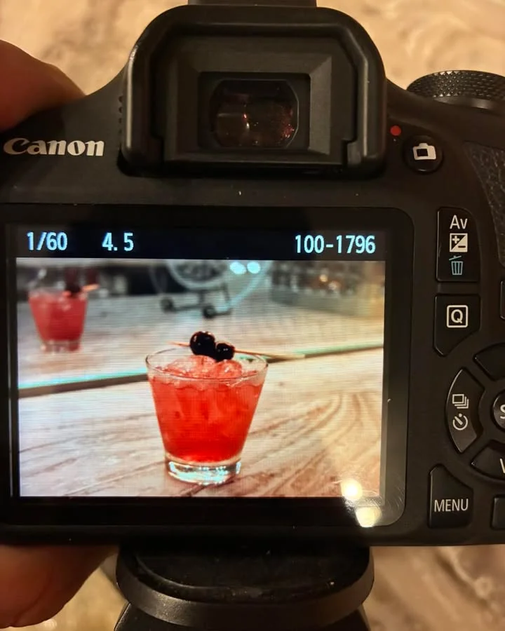 A close-up shot of a cocktail glass filled with a vibrant red drink, garnished with dark berries, captured through a camera viewfinder.