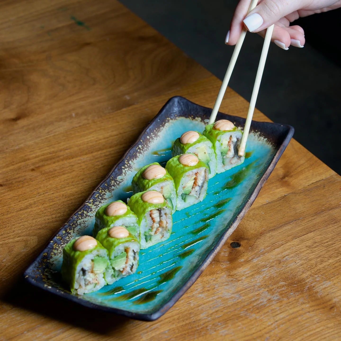 Sushi rolls wrapped in bright green soy paper or avocado, topped with spicy mayo and served on a turquoise rectangular plate being picked up with chopsticks.