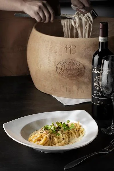 A plate of spaghetti served with sauce, garnished with microgreens, alongside a large wheel of cheese and a bottle of red wine.