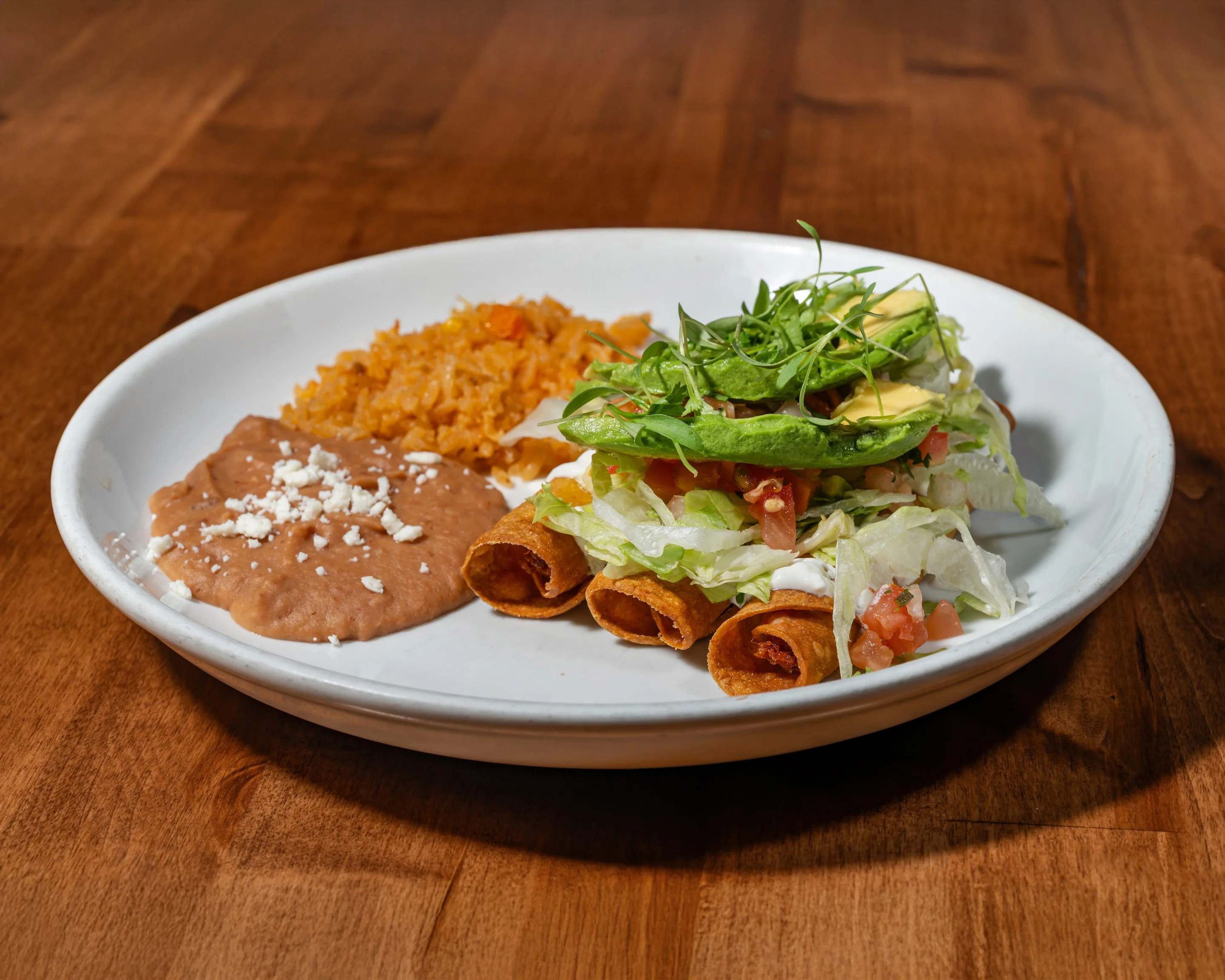 A plate featuring rolled tortillas filled with meat, topped with lettuce, tomatoes, avocado, and a sprinkle of cheese. Accompanied by refried beans and orange rice.