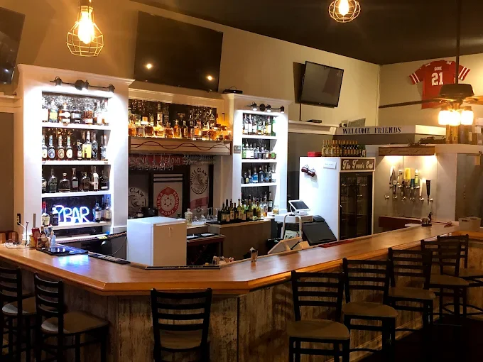 An inviting bar area featuring a wooden counter with several high-backed chairs. Behind the bar, shelves are stocked with various bottles of alcohol, and there are two large televisions mounted on the wall.