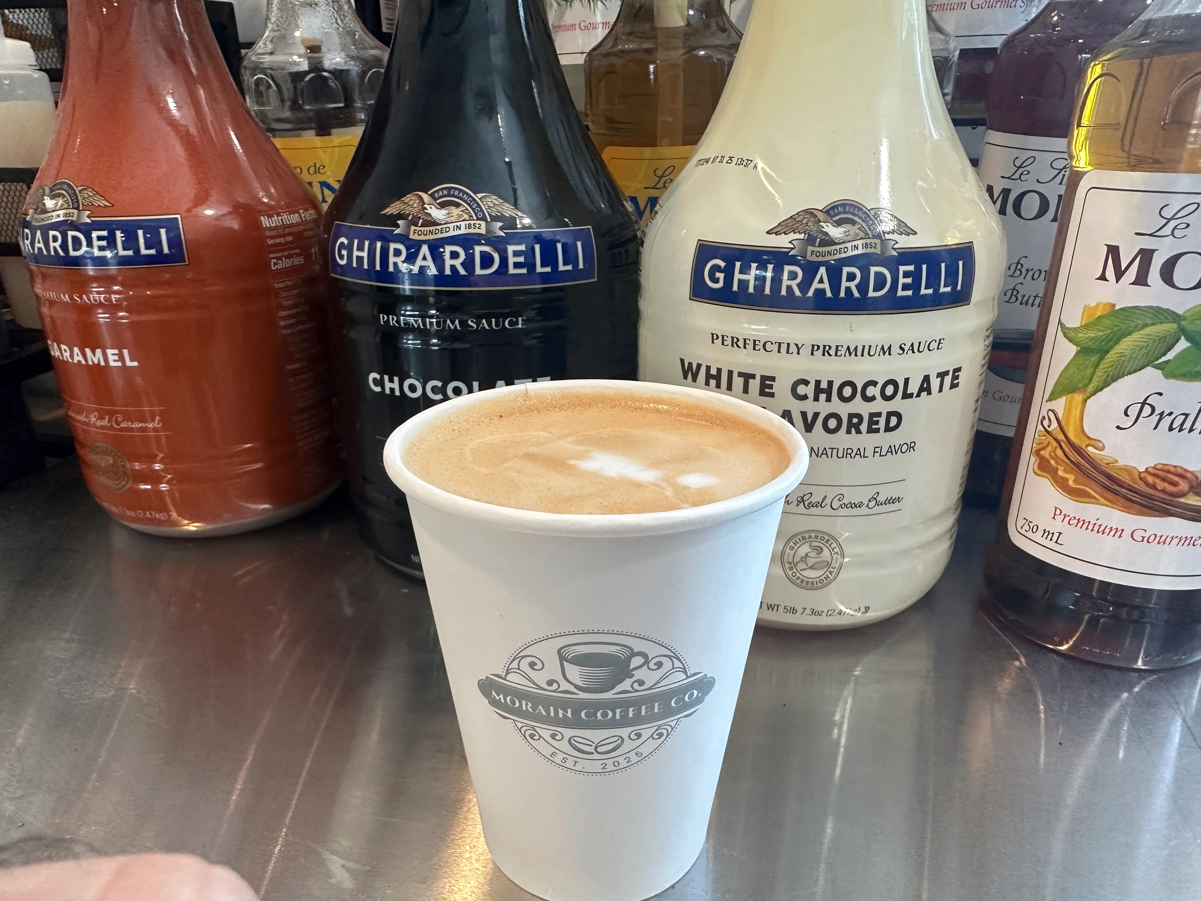 Coffee cup with latte art beside Ghirardelli sauces