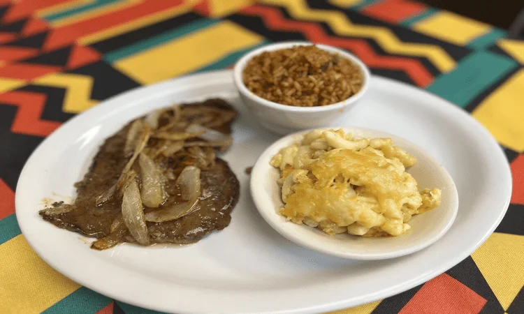 A plate with sliced beef smothered in onions, alongside macaroni and cheese and seasoned rice in separate bowls, arranged on a bright patterned tablecloth.
