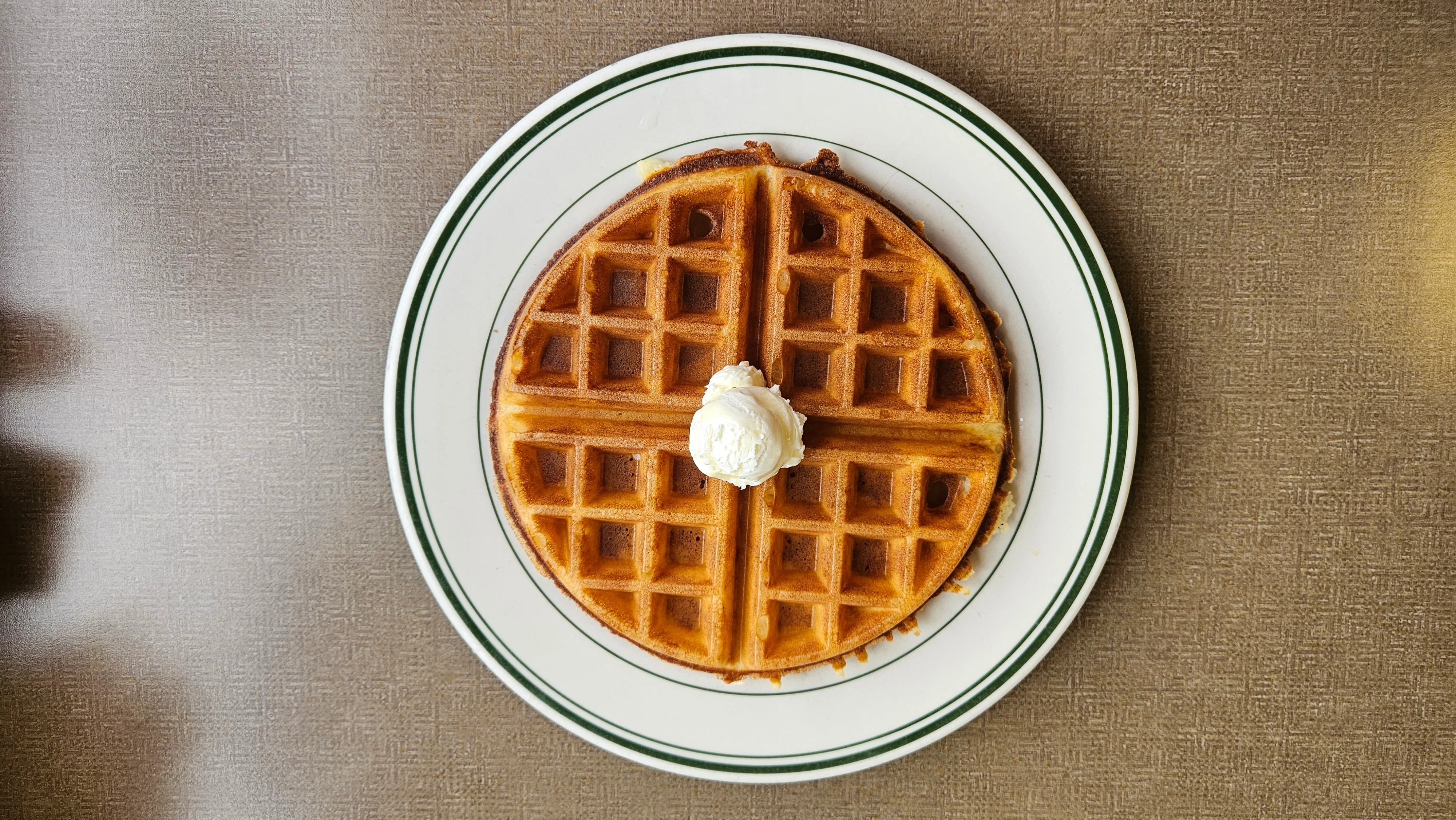 A golden brown waffle topped with a dollop of whipped cream, served on a white plate with green trim.