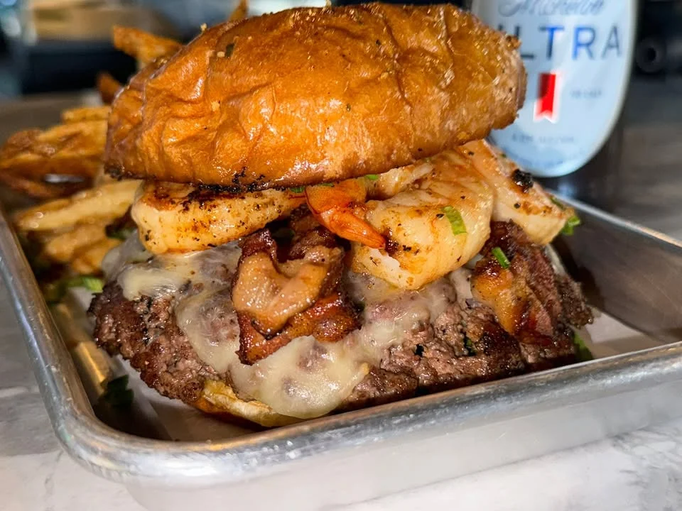 A gourmet burger topped with shrimp and bacon, served with a side of fries in a metal tray.