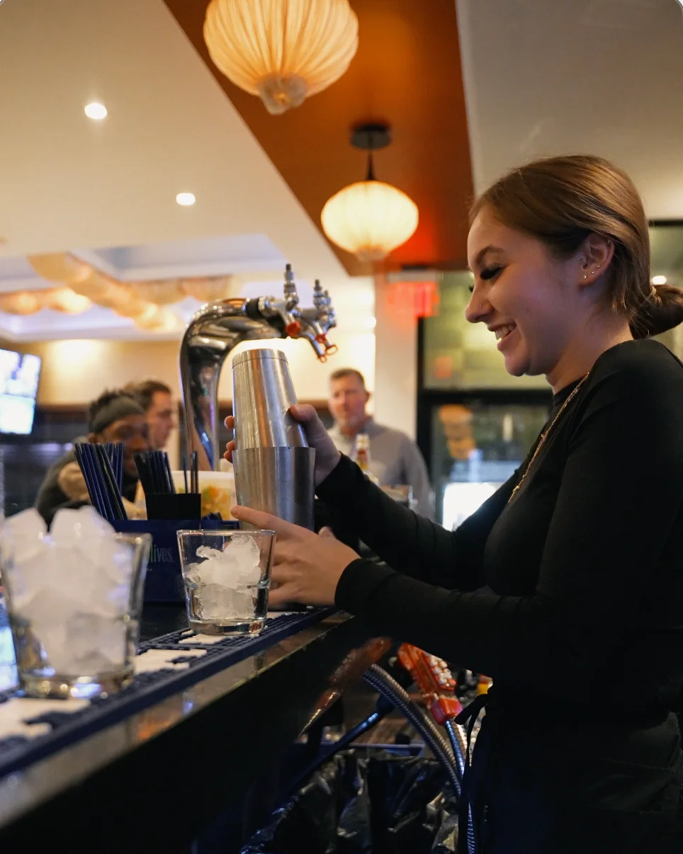 Bartender shaking a cocktail at a lively bar with patrons in the background.
