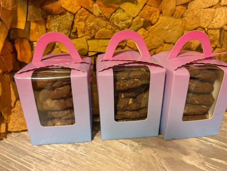 Three clear boxes containing stacked cookies, showcasing a variety of cookie types. The boxes have pastel-colored bottoms and pink handles.