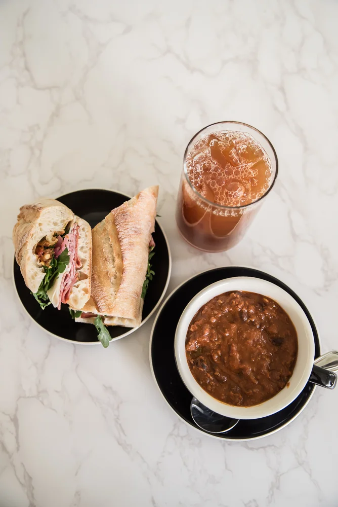 A sandwich with greens and meat on a black plate, a cup of chili soup on a saucer, and a glass of iced tea are set on a marble surface. Cozy and appetizing.