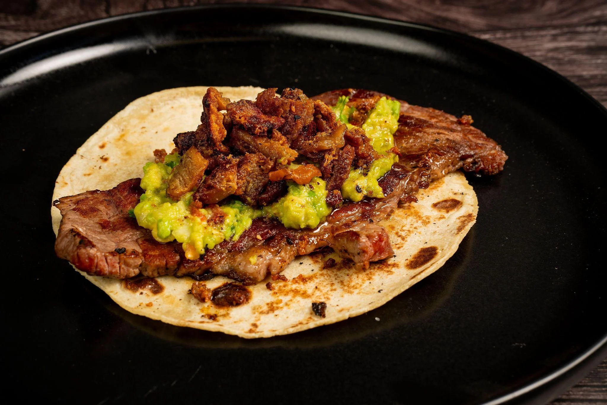 A taco filled with grilled beef and topped with crispy pork and a dollop of guacamole, served on a black plate.