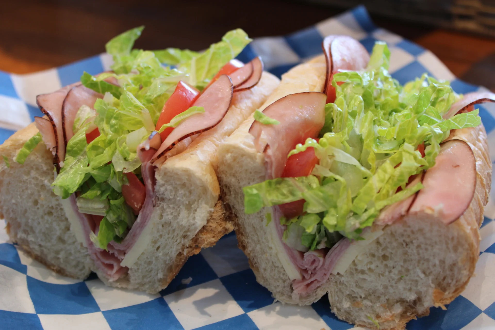 A sub sandwich filled with layers of sliced meats, lettuce, and tomatoes, presented on a blue-checkered paper.
