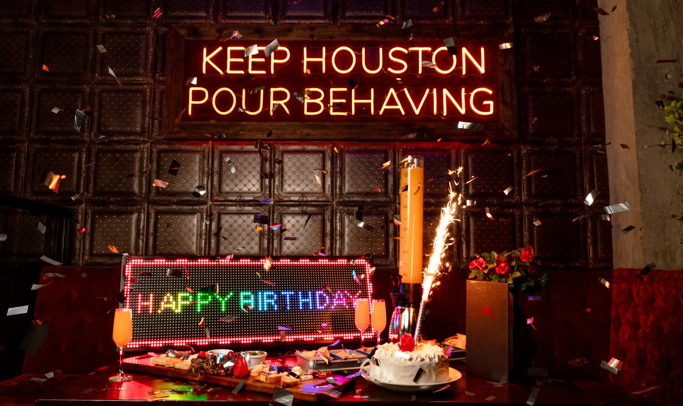 Birthday celebration setup at Pour Behavior in Houston, featuring confetti, sparklers, champagne, cake, and a neon sign that reads ‘Keep Houston Pour Behaving.