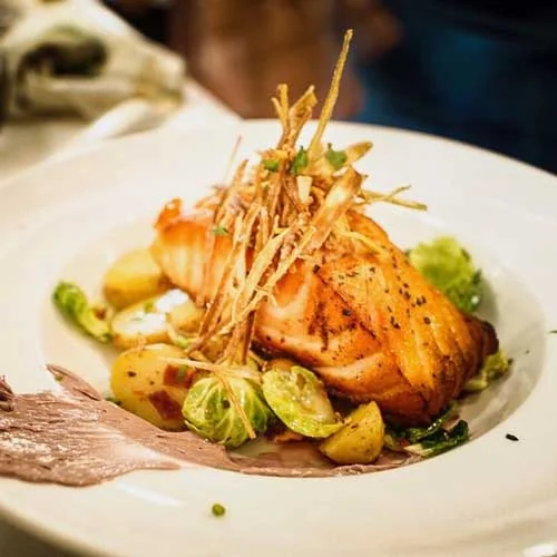White plate with cooked salmon, brussel sprouts, potatoes, strips of tortilla, and a brown sauce smeared on the plate