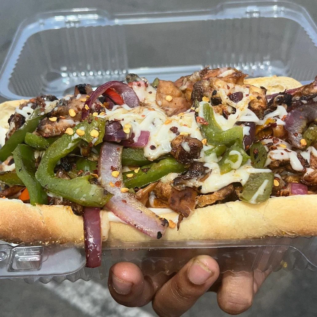 A loaded sandwich featuring grilled meat, colorful bell peppers, and melted cheese on a toasted bun, served in a takeout container.