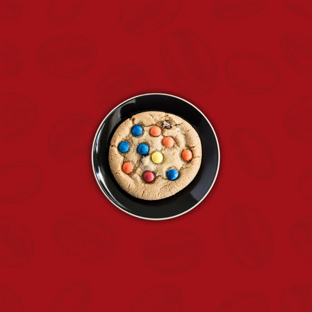 A cookie with colorful candies sits on a black plate against a rich red background. The playful candies add a touch of fun to the inviting scene.