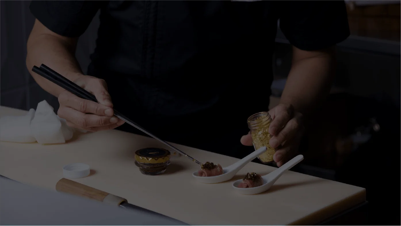 A chef wearing a branded Ke-uH uniform uses long metal chopsticks to delicately place a combination of caviar and edible gold flakes onto small, rolled appetizers resting in white ceramic spoons.