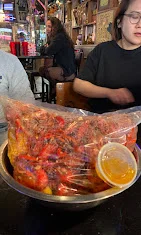 A large bowl filled with crawfish, served in a bag with a cup of dipping sauce. The setting is casual with a bar in the background.