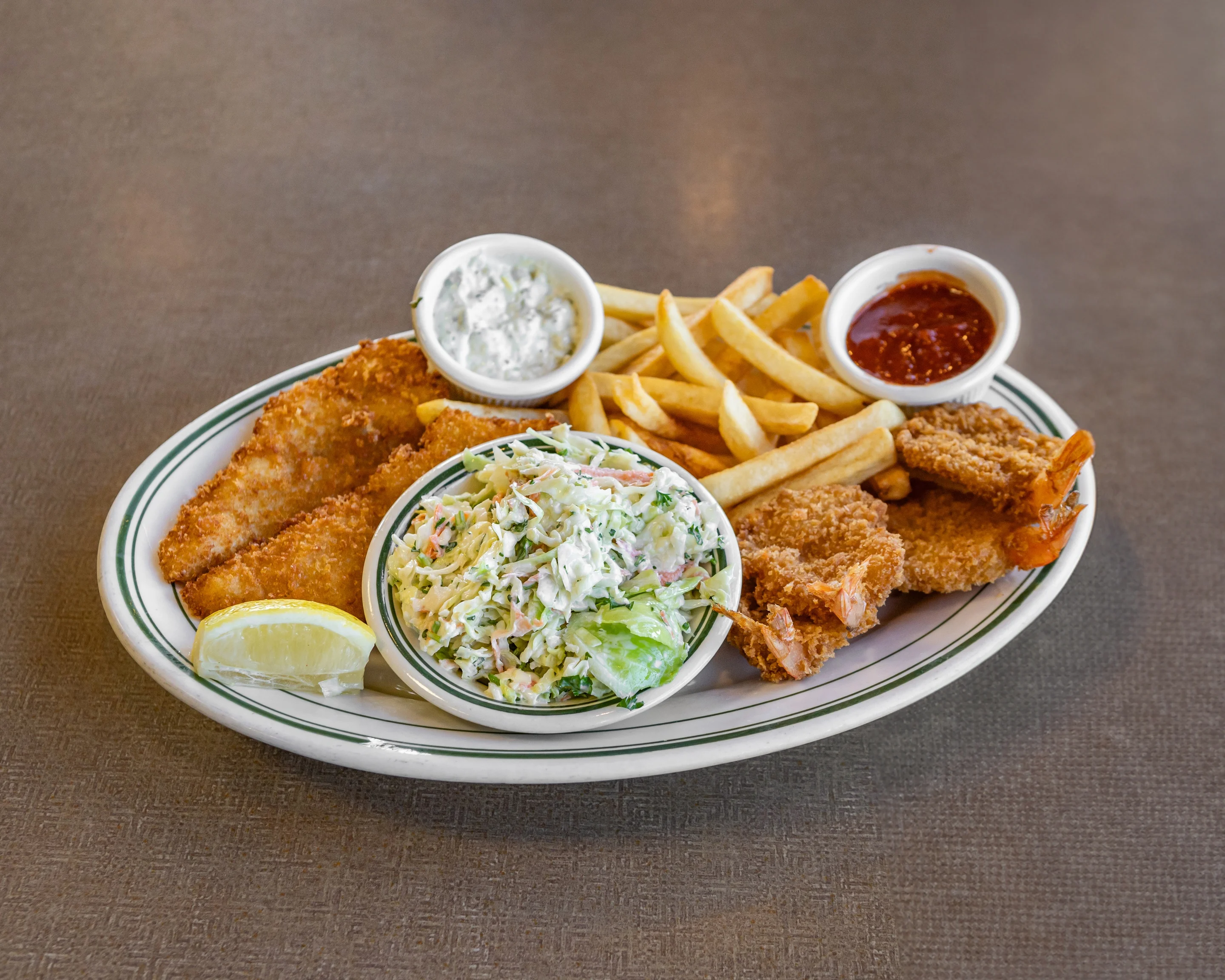 A seafood platter with fried fish fillets, shrimp, french fries, coleslaw, and dipping sauces, garnished with a lemon wedge.